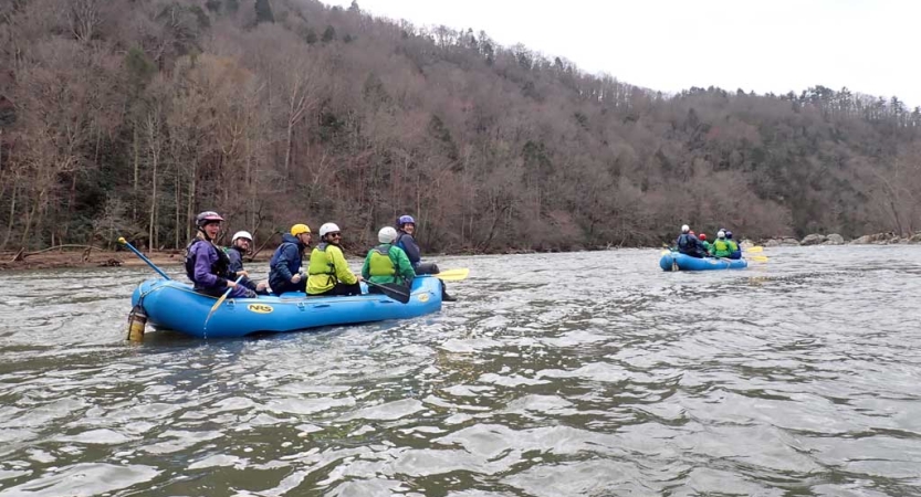 Two rafts are navigated by people wearing safety gear on a calm river. 